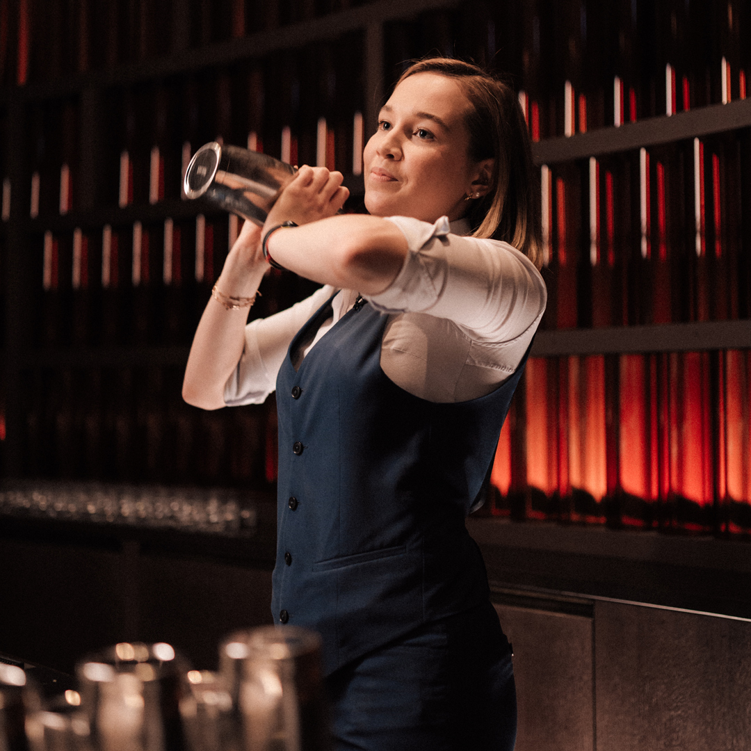 Junge blonde Marriott Barkeeperin mit weißem Hemd und blauer Weste mixt Cocktail in einem Shaker an einer Hotelbar