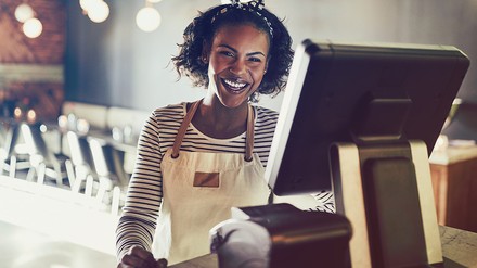 Schwarze lächelnde Frau, die eine weiße Schürze trägt und hinter einer Restaurantkasse steht