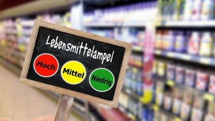 Eine Hand hält in einem Supermarkt ein Schild hoch auf dem die Lebensmittelampel zu sehen ist. Rot für hoch, gelb für mittel und grün für niedrig