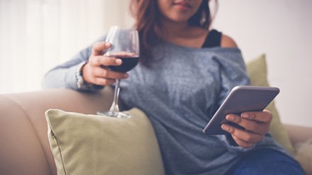 Junge Frau sitzt auf der Couch, trinkt Wein und hältein Smartphone in der Hand