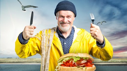 Ein Fischer hält Messer und Gabel hoch um ein vor ihm liegendes Fischbrötchen zu essen