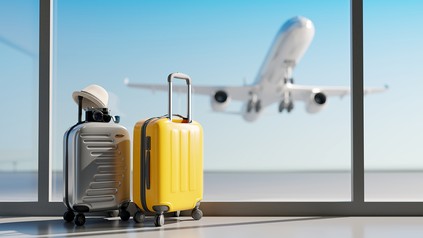 Zwei Koffer vor einer Glasfassade am Flughafen, im Hintergrund ein startendes Flugzeug