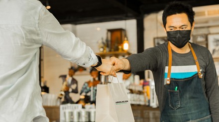 Ein Mann mit Mund-Nasen-Maske gibt einem Kunden eine Papiertüte mit To-go-Speisen