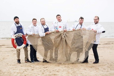 Sechs Köche halten gemeinsam ein Fischernetz an einem Strand