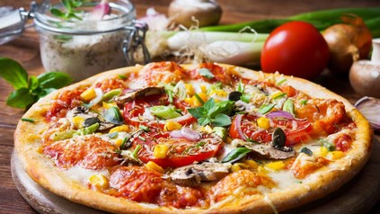 Eine frisch zubereitete und reichlich belegte Pizza und die Zutaten im Hintergrund