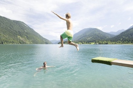 Ein Mann springt von einem Sprungbrett in einen See in dem bereits ein anderer Mann schwimmt