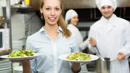 Eine Serviekraft mit zwei Tellern Salat, im Hintergrund zwei Köche bei der Arbeit