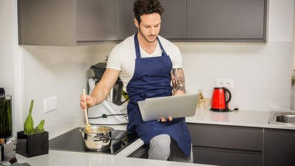 Ein junger Mann mit Schürze sitzt auf der Arbeitsfläche seiner Küche, rührt in einem Kopf und blickt dabei auf den Laptop, den er auf seinen Knien hält
