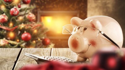 Ein weihnachtlich eingerichtetes Zimmer mit einem Weihnachtsbaum, davor sitzt ein Sparschein mit Brille auf einem Tisch vor einem Taschenrechner