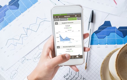 Eine Person hält ein Smartphone auf dem die App Gastronovi Cockpit aufgerufen ist, darunter liegen mehrere Aufzeichnungen und Grafiken