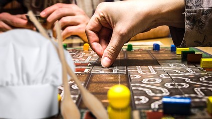 Zwei Personen knobeln an einem Brettspiel, davor sind eine Kochmütze und zwei Kochlöffel abgebildet