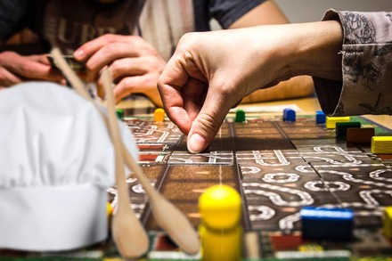 Zwei Personen knobeln an einem Brettspiel, davor sind eine Kochmütze und zwei Kochlöffel abgebildet