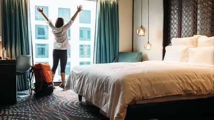 Frau streckt vor dem Fenster eines Hotelzimmers die Arme in die Luft