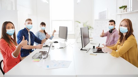 Sechs junge Leute in einem Meeting, tragen Mund-Nasen-Masken und sitzen vor PC-Bildschirmen und Laptops, halten Daumen nach oben