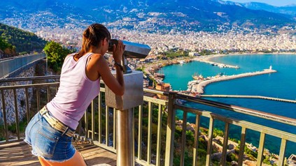 Eine junge Frau blickt durch ein Fernglas auf einen kleinen Hafen in einer türkischen Stadt