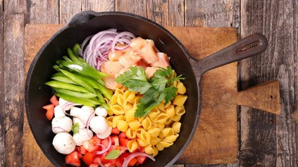 In einer Pfanne befinden sich Nudeln, Fleisch, geschnittene Zwiebeln, Paprika, Pilze und Tomaten mit einem Bund Petersilie