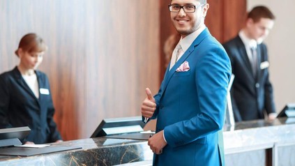 Ein Geschäftsmann steht vor einer Hotelrezeption und macht lächelnd die Daumen-Hoch-Geste