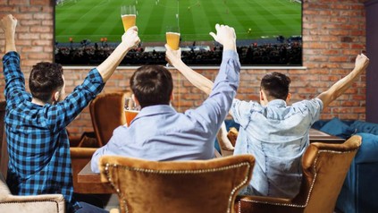Drei Junge Männer jubeln mit Bier in der Hand vor einem großen Fernseher auf dem ein Fußballspiel läuft