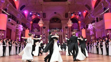 Männer in schwarzen Fracks und Frauen in weißen Kleidern tanzen im Kreis, umringt von Personen in derselben Kleidung