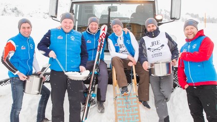 Sechs Köche mit Pfannen und Töpfen im Schnee vor einer Schneeraupe