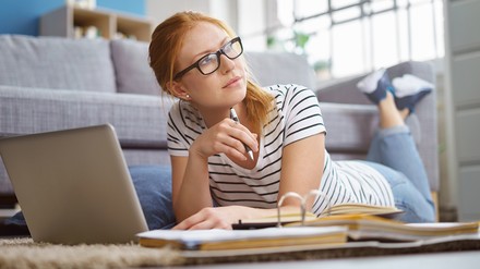 Junge Studentin oder Auszubildende lernt vor dem Laptop auf dem Fußboden, drum herum liegen Ordner und Bücher