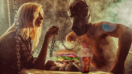 Eine Frau sitzt vor einem Burger und einem Getränk und hält einen Mann mit Tiermaske an einer Kette