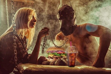 Eine Frau sitzt vor einem Burger und einem Getränk und hält einen Mann mit Tiermaske an einer Kette