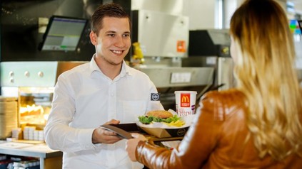 Ein freundlicher McDonald's Mitarbeiter händigt einer Kundin ein Tablett mit Burger und Getränk aus