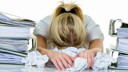 Eine junge Frau verzweifelt im Büro zwischen vielen Aktenordnern und zerknülltem Papier.