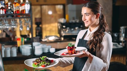 Junge Servicekraft trägt lächelnd zwei Teller zum Tisch