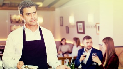 Ein genervter Kellner hält einen kleinen Teller mit ein paar Euromünzen in der Hand und hält die andere Hand erwartungsvoll auf