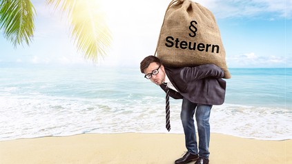 Ein Mann am Strand trägt einen Sack, auf dem Steuern steht.