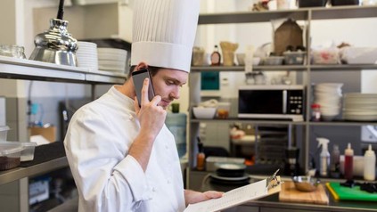 Ein Koch steht mit einem Klemmbrett in der Küche und telefoniert mit seinem Smartphone