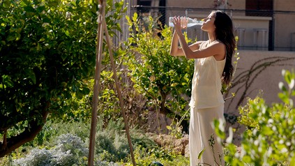 Eine Frau steht bei Sonnenschein in einem Garten und trinkt Wasser aus einer Glasflasche.