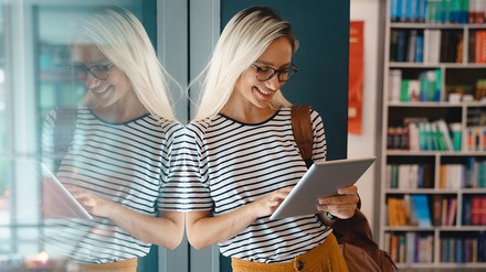 Studentin in einer Bibliothek schaut lächelnd in ein Tablet
