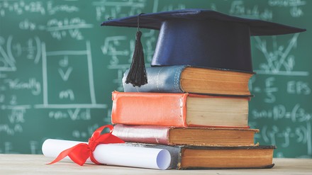 Dicke Lehrbücher liegen auf einem Tisch, darauf liegt ein Stipendiatenhut. Im Hintergrund steht eine vollgeschriebene Tafel.