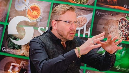 Frank Simmeth in dunklem Hemd gestikuliert vor einer Fotowand