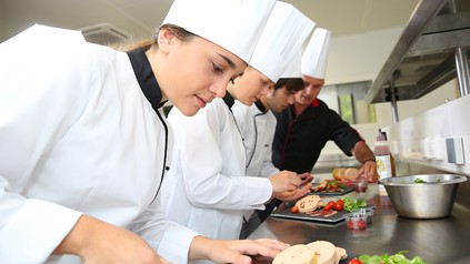 Köche stehen in einer Küche und bereiten ein Gericht vor. Ein Ausbilder gibt Anweisungen.