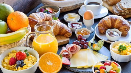 Ein reich gedeckter Frühstückstisch mit Wurst- und Käseplatte, Brötchen, Ei, Müsli, Obst und Orangensaft.