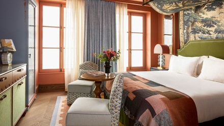 Ein stilvolles Hotelzimmer im Le Grand Mazarin in Paris, das mit seiner warmen Farbpalette und raffinierten Einrichtung Eleganz ausstrahlt. Das Zimmer verfügt über ein großes Bett mit einer grünen Kopflehne und einer geometrisch gemusterten Decke in Erdtönen. Neben dem Bett befindet sich ein kleiner Sitzbereich mit einem runden Tisch und einer Vase mit frischen Blumen. Hintergrundbild und Vorhänge ergänzen das anspruchsvolle Design, während natürliches Licht durch die französischen Fenster strömt und das Zimmer in ein einladendes Ambiente taucht.