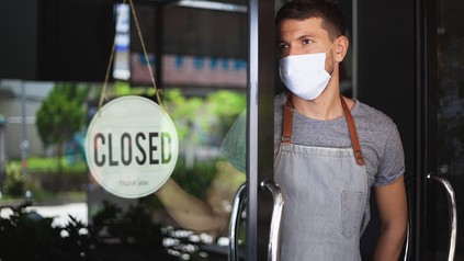Kellner mit Mund-Nasen-Maske hängt ein "Closed"-Schild ins Fenster eines Restaurants oder einer Bar