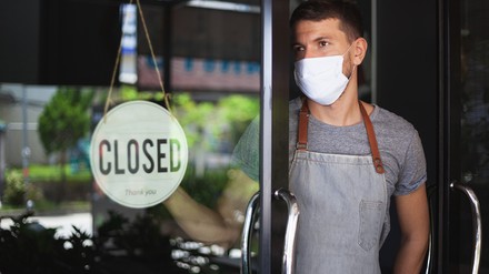 Kellner mit Mund-Nasen-Maske hängt ein „Closed“-Schild ins Fenster eines Restaurants oder einer Bar