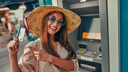 Eine junge Frau mit Sommerhut steht vor einem Geldautomat und hebt ihre Kreditkarte in die Kamera.
