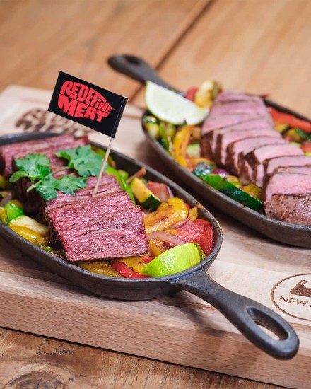 Zwei Gusseiserne Servierpfannen mit Gemüse und künstlichem Fleisch in der einen und echtem Fleisch in der anderen stehen nebeneinander auf einem Holzbrett