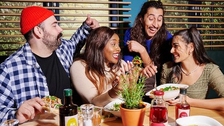 Vier Freunde, zwei Männer und zwei Frauen, verschiedener Ethnien sitzen lachend mit veganen Gerichten an einem Restauranttisch auf dem Getränke und ein Topf mit Kräutern steht