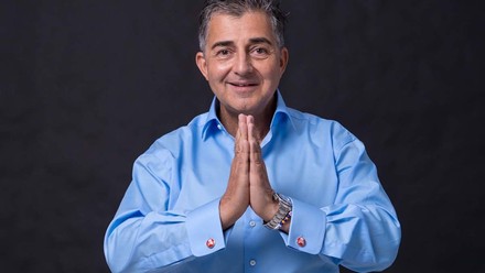 Pierre Nierhaus sitzt im blauen Hemd mit gefalteten Händen vor einer dunklen Wand