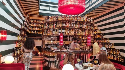 Ein lebendiges Restaurantinterieur mit schwarz-weiß gestreiften Wänden, einer großen, runden Bar mit einer beeindruckenden Auswahl an Spirituosen und einem auffälligen roten Kronleuchter. Gäste genießen Getränke und Unterhaltungen an der Bar.