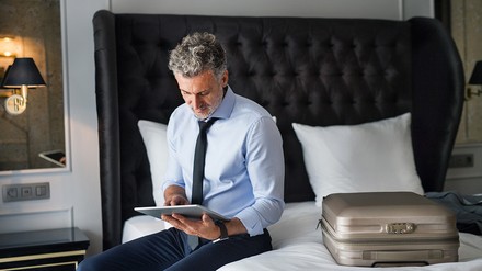 Mann sitzt mit Koffer und Tablet in der Hand auf einem Hotelbett