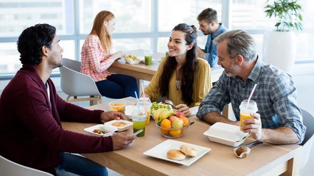 Fünf Menschen sitzen an unterschiedlichen Tischen in einer Kantine und essen gemeinsam.
