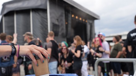 Junge Menschen auf einem Festival im Hintergrund, im Vordergrund wird ein Becher in einen Müllkorb geworfen
